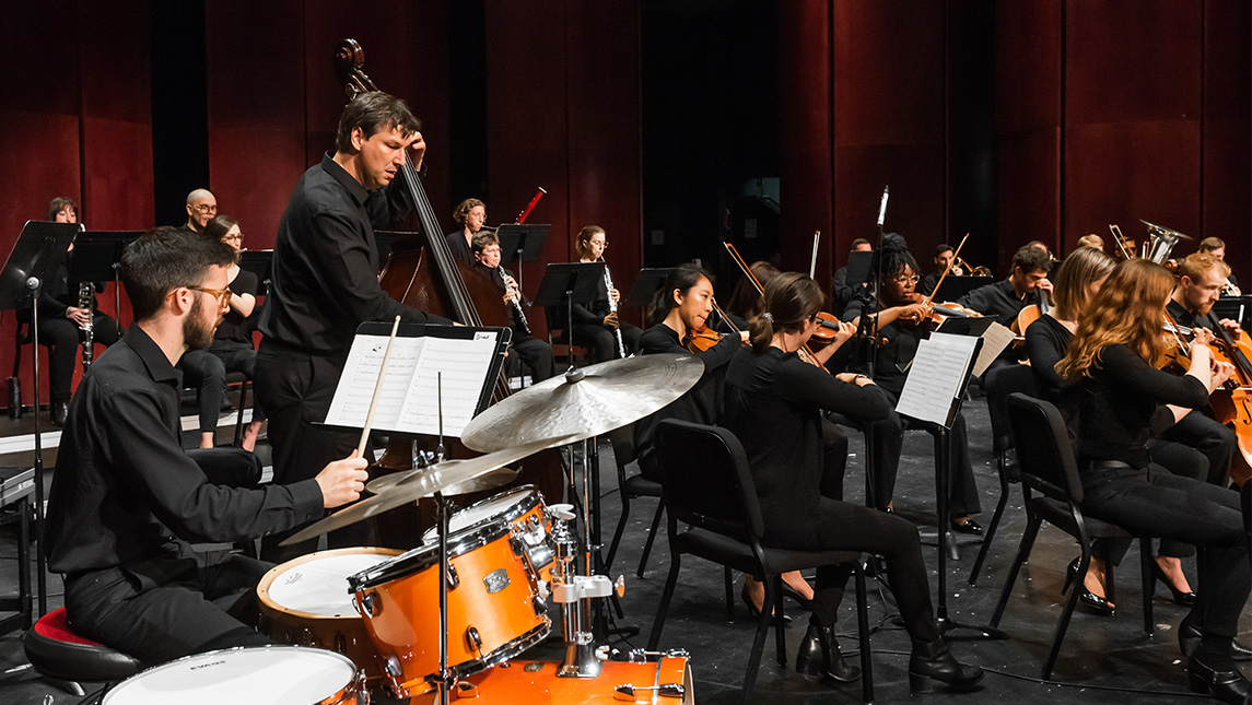UNCG students play drums and strings in an orchestra.