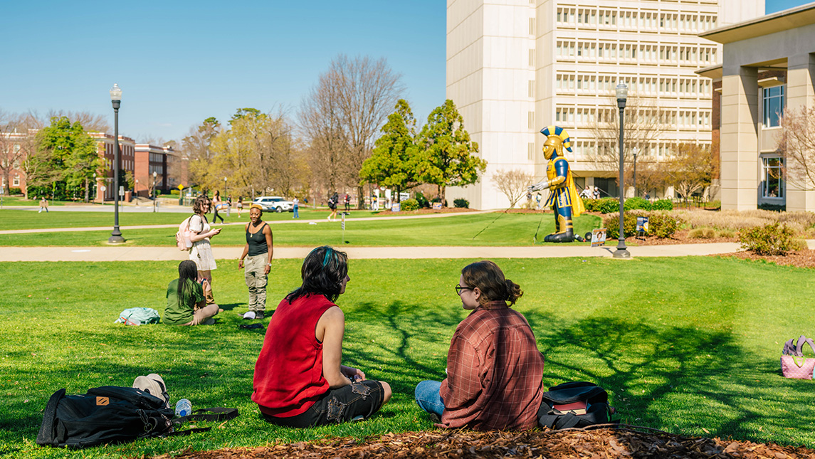 Students sit on the lawn outside the UNCG EUC with an inflatable Spiro mascot in the background.