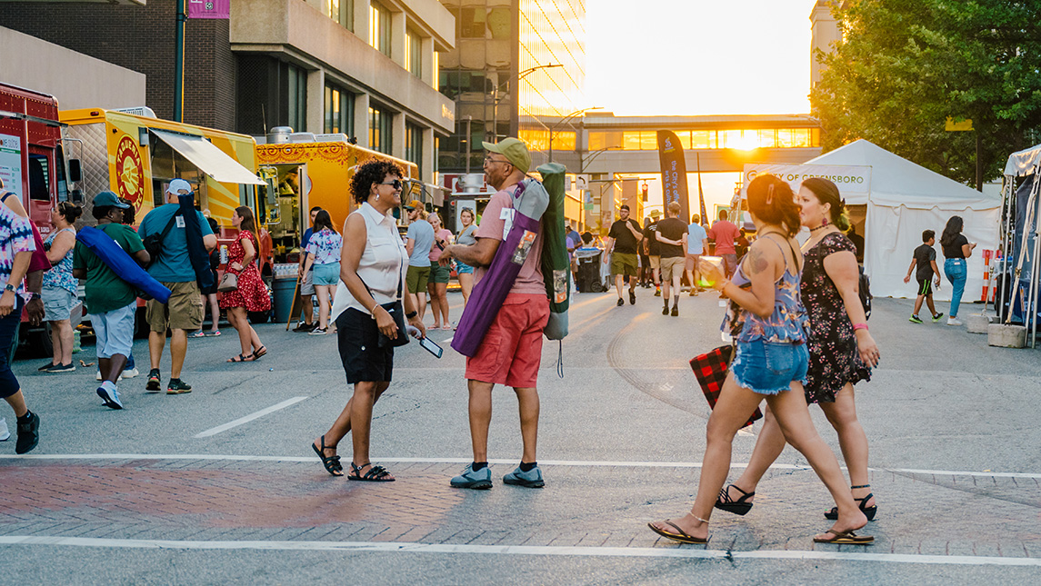 People walk through downtown streets with sun setting behind the buildings and food trucks in the background.