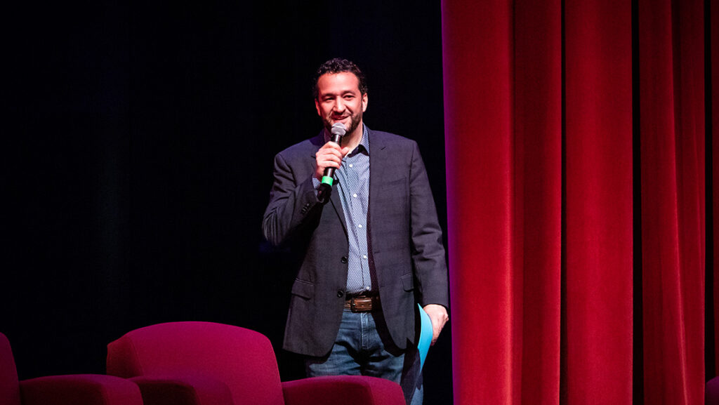 Man stands on a stage with a microphone and a red curtain behind him.