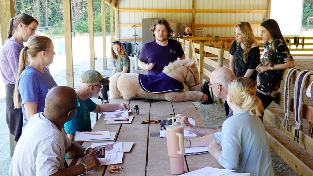 A large group of people gather around a stuffed animal horse inside a stable.