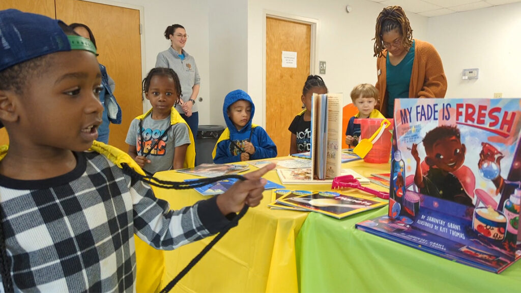 A boy looks at a picture book with UNCG alumna Dr. Tyla Ricks in the background.