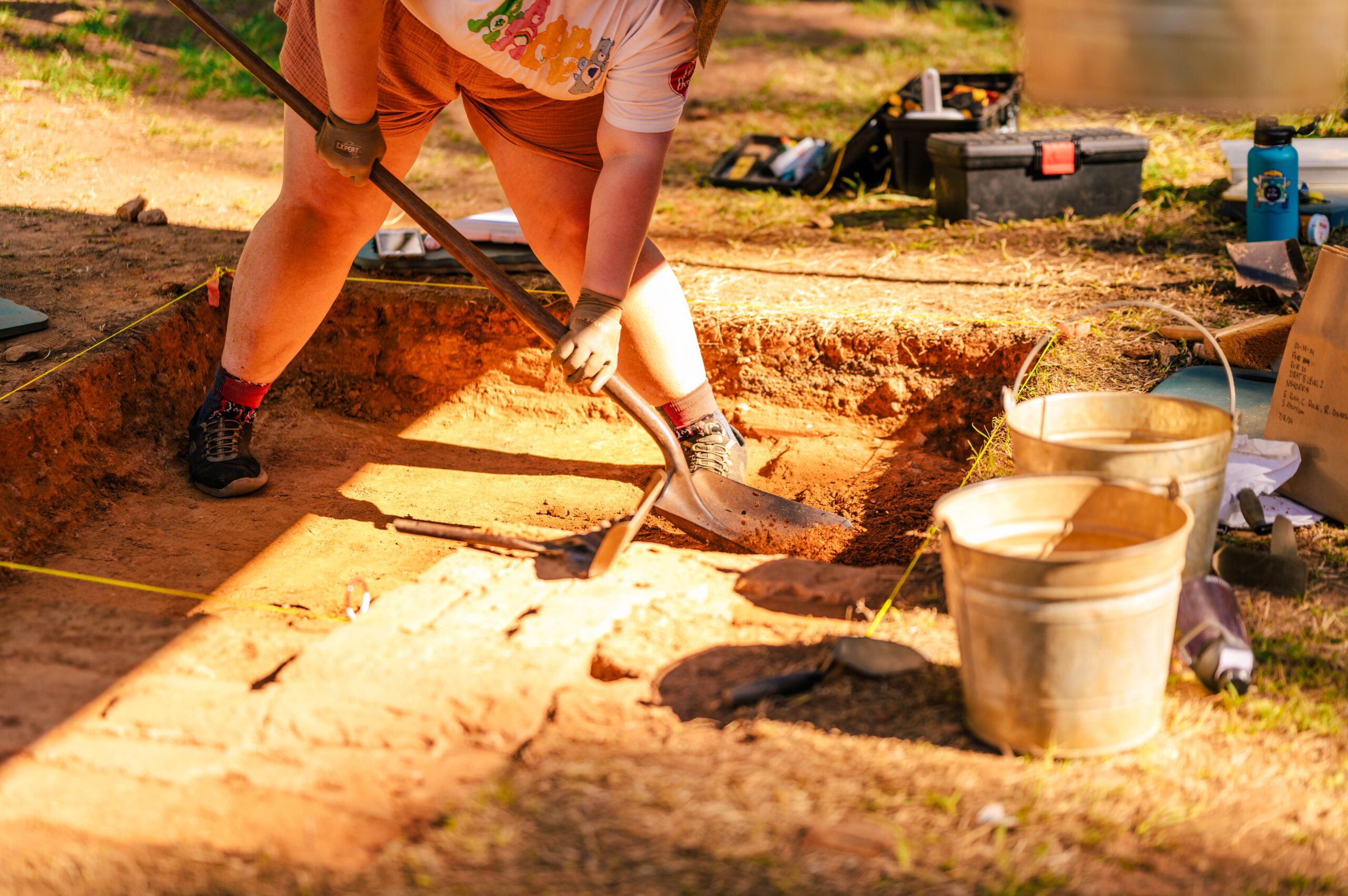 Close-up of person digging a measured plot in the ground with tools and buckets around the hole.