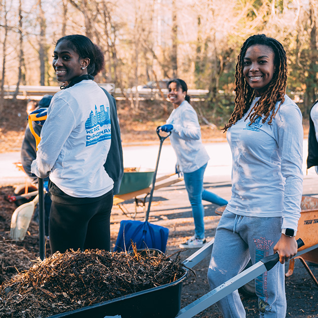 Girl with a wheelbarrow full of mulch smiles at the camera with 2 friends with shovels behind her.