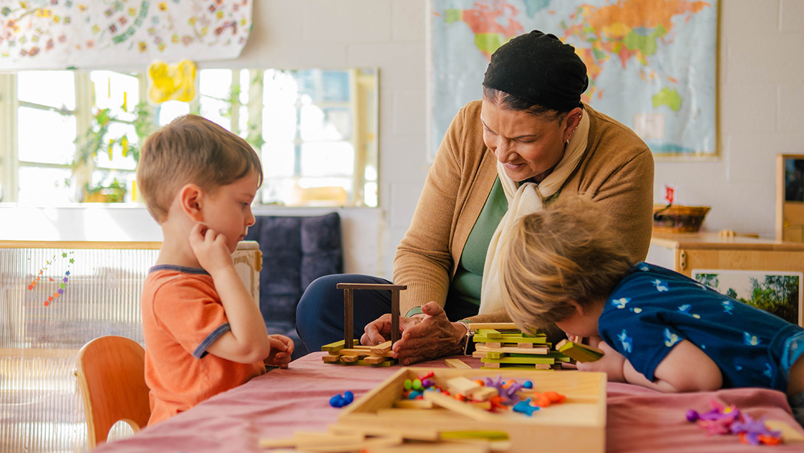 A woman helps two small boys build with blocks.