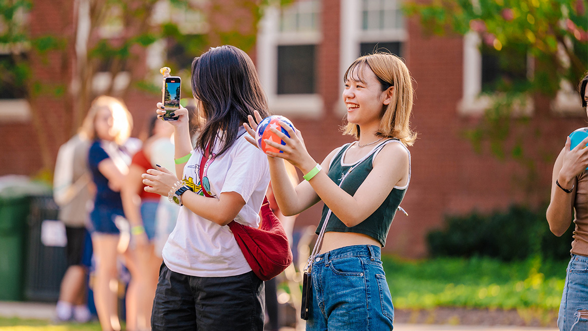 Two girls throw a ball and take video with their phone at UNCG.