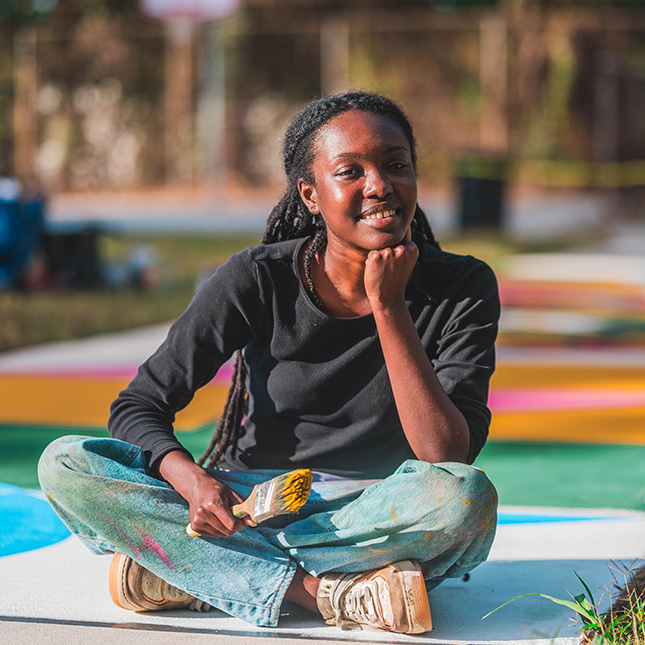 Girl sits cross-legged on a painted park sidewalk with a paint brush.