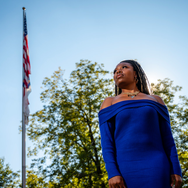 Woman in blue dress looks up towards a flag pole with the American & NC flags raised.