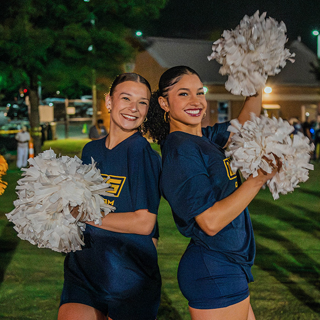 Two dance team members with pompoms pose on a dark athletic field.