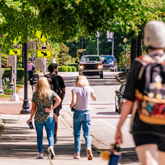 Students walking away from camera on a sidewalk with beside a road with oncoming cars.