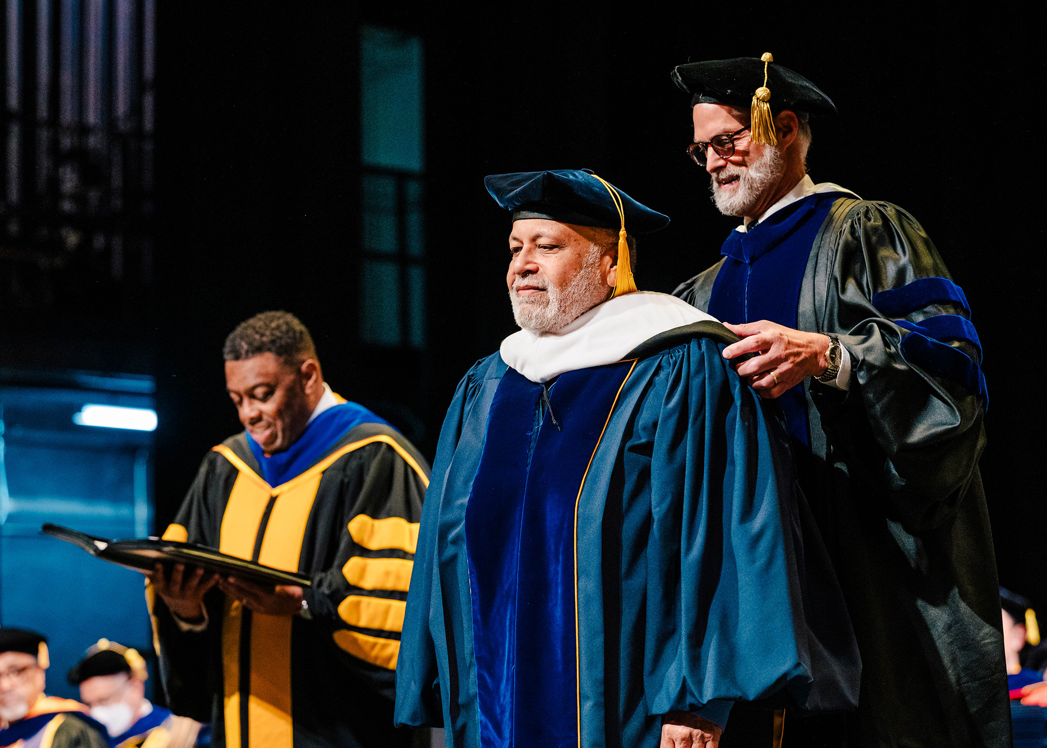 Faculty member in gown and cap places a doctoral hood on the shoulders of a man with the UNCG Chancellor looking on.