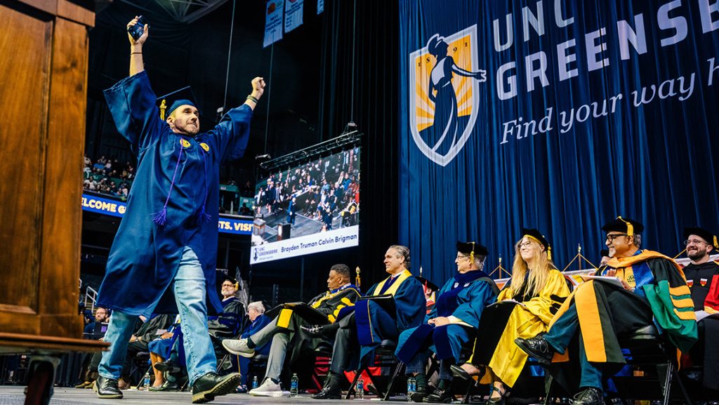UNCG graduate crosses the stage in cap and gown with hands outstretched as faculty look on.