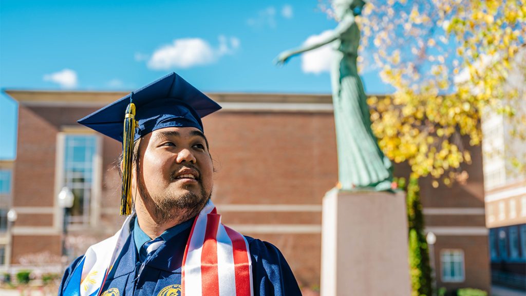 Elijah Resuello in his cap and gown with UNCG Minerva statue in the background.