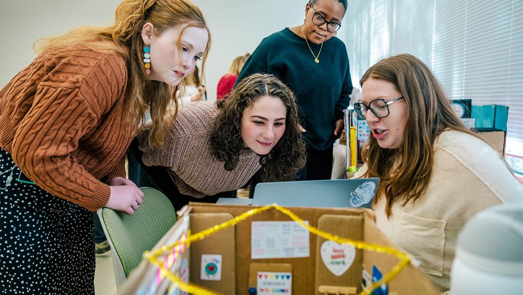 UNCG students gather around a cardboard model of a classroom.