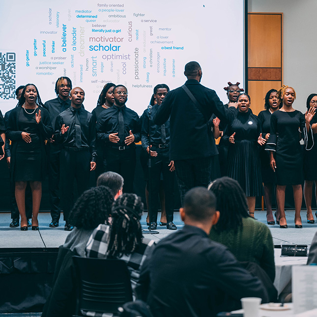Crowd sits at tables watching a choir sing on a stage.