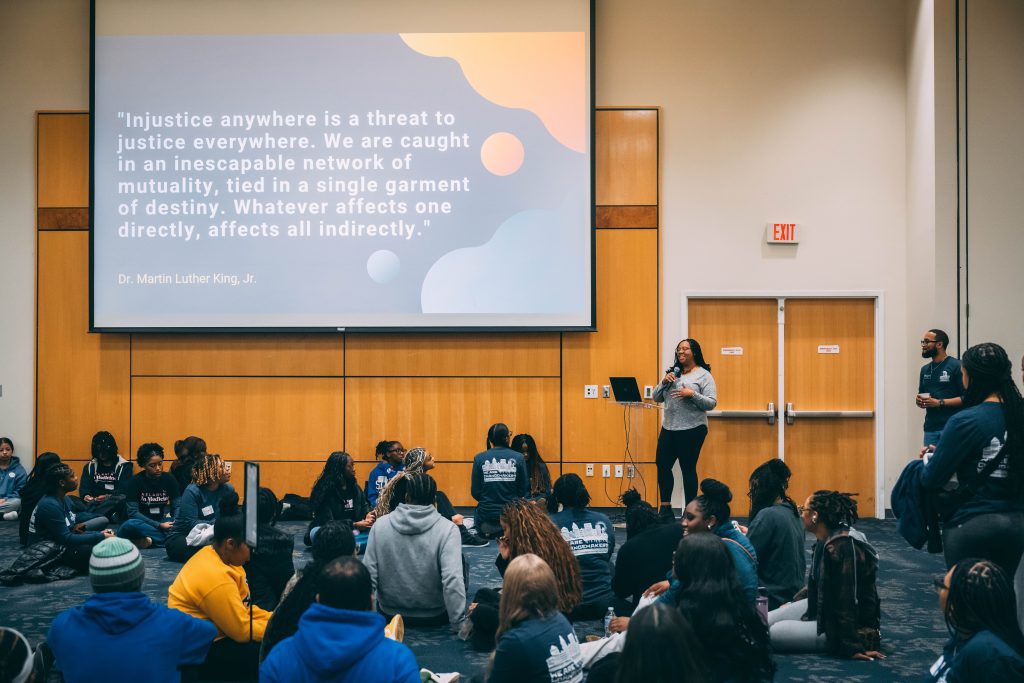 Group of students sit on the floor of the EUC ballroom and listen to a speaker with a MLK quote on the screen above.