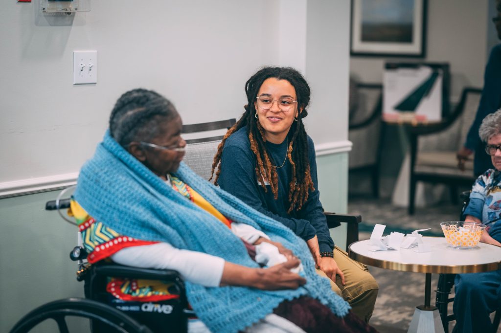 A student talks to an elderly woman sitting in a wheelchair in a lobby of a retirement facility.