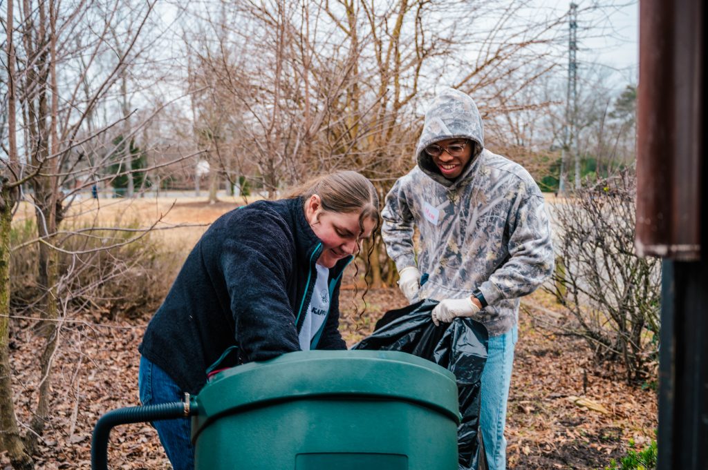 Two students pictured outdoors in a garden loading a lawn trash bin.