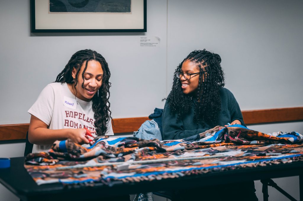 Two women work at a table cutting fabric and chatting together.