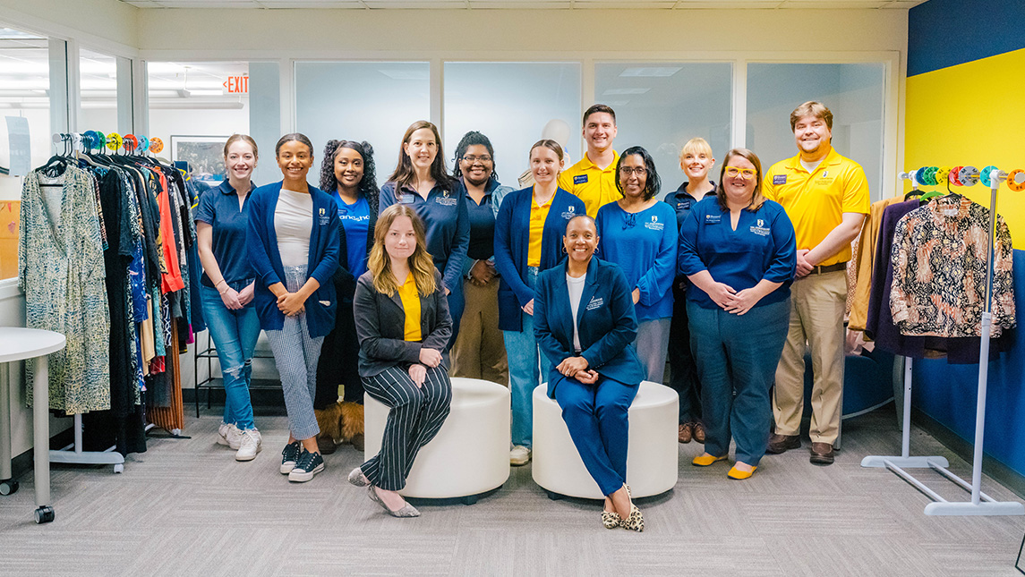 Group in UNCG staff shirts pose in the CPD suite with racks of clothes on either side of them.