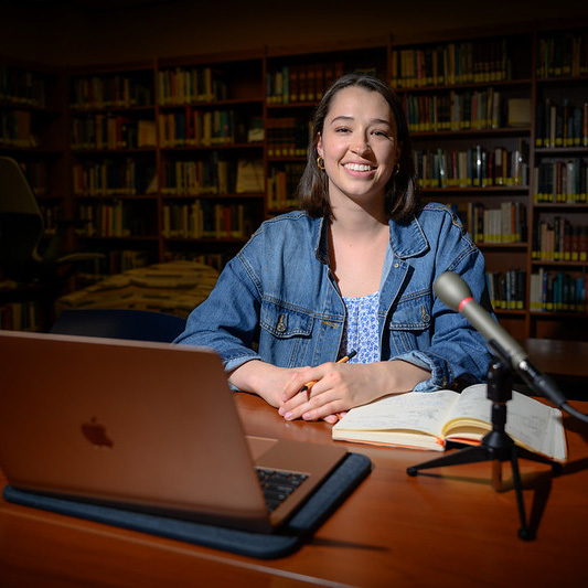 A student sits at a table in a library with a laptop, notebook, and microphone, studying or recording work.