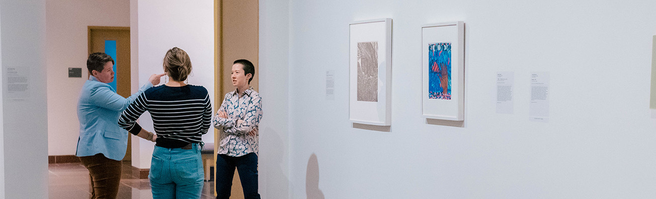 Three professionally dressed people chat in an art museum gallery while one points to artwork.