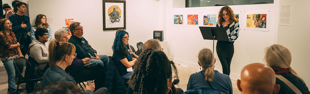 Woman reads at a podium to a group of people sitting in chairs in an art gallery.