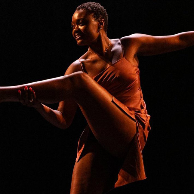 Dancer on a dark stage smiling as she lifts her leg and stretches.