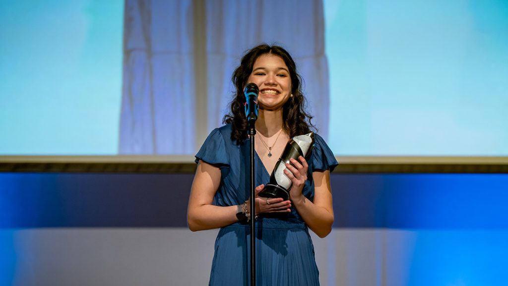 Woman accepts an award on a stage and smiles broadly behind a microphone.