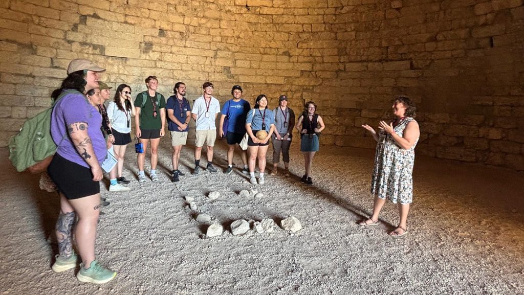 A group of students stand in a semi circle in listening to their instructor in a ruin on the Greek Island of Kea