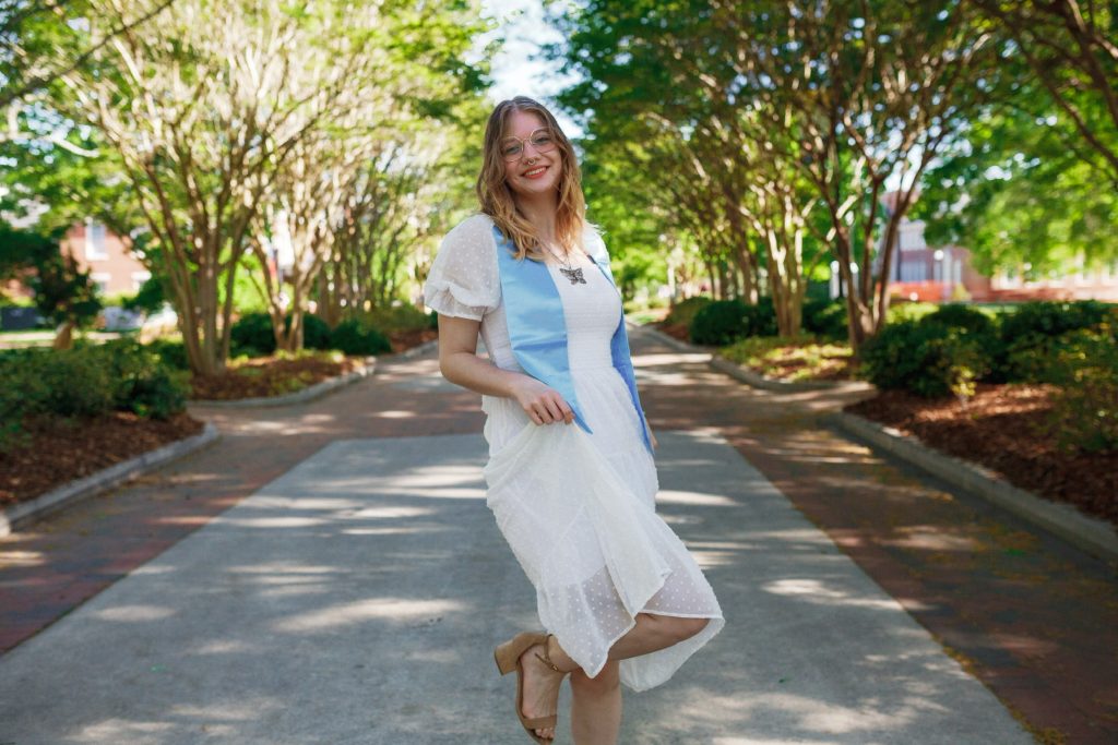 Alyssa Causey wears a white dress and a light blue stole while standing on a tree-lined sidewalk