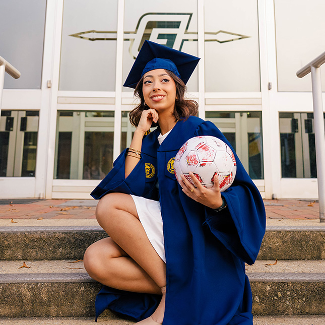Grad in cap and gown sits on steps of Coleman gym holding a soccer ball.