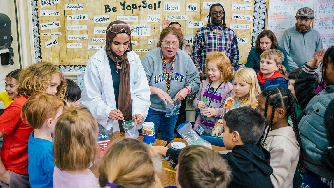 A group of students gather around a college student in a lab coat doing a science experiment in an elementary classroom with teachers and parents looking on.
