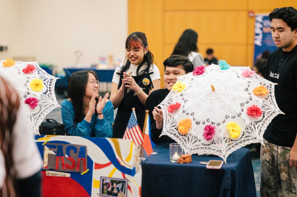 Three students sitting behind a stable at a cultural fair. International flags and decorative umbrellas rest on the table.