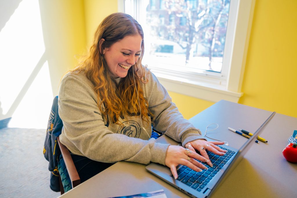 A student sit behind a desk, working on a a laptop and smiling.