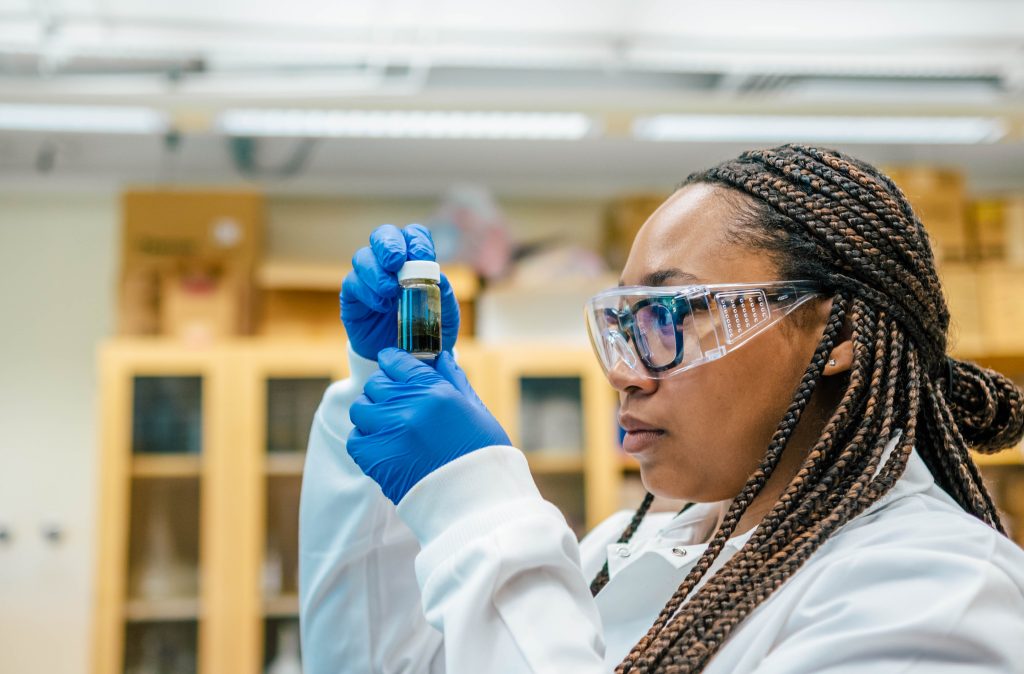 A chemistry student wearing a lab coat, gloves, and protective glasses holds and observes specimen vial in a laboratory setting.