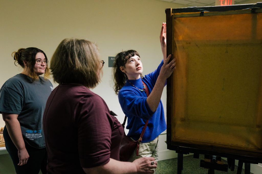 A student careful holds an old piece of artwork, making sure it's wrapping stays in place as a fellow student and instructor look on.