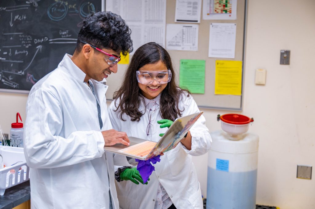 Two chemistry students student together in a science classroom, smiling looking at a laptop. Both are wearing lab coats and goggles.