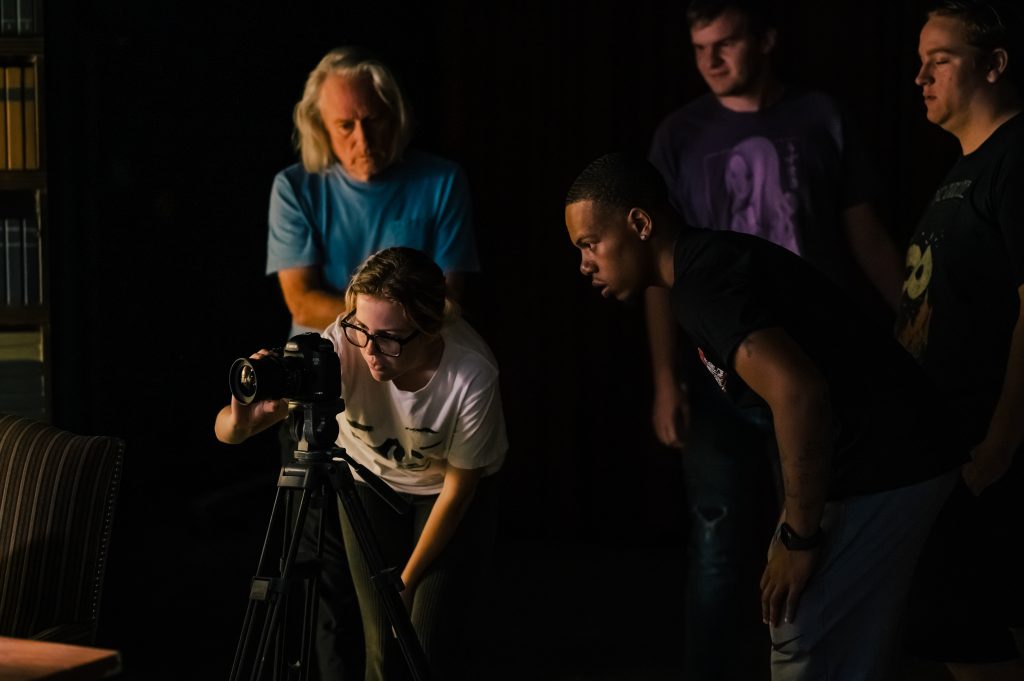 A group of students an instructors gather around a video camera. One student works to adjust the zoom.
