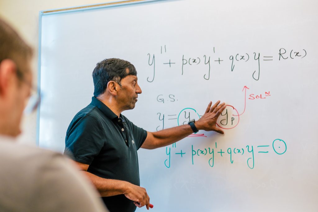 A professor standing at the white board pointing to and explaining an equation