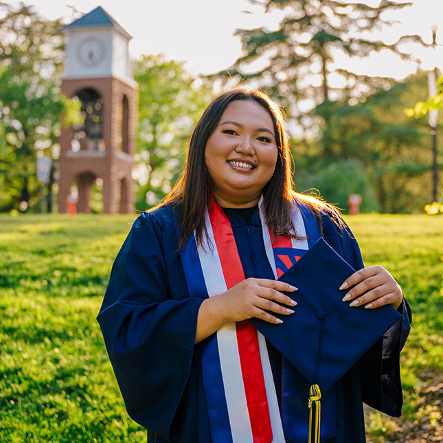Graduate in gown holds cap and poses in front of UNCG's clock tower.