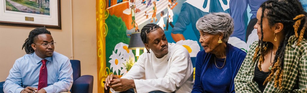 UNCG students sit with Joanne Smart Drane in front of a mural painting of her.