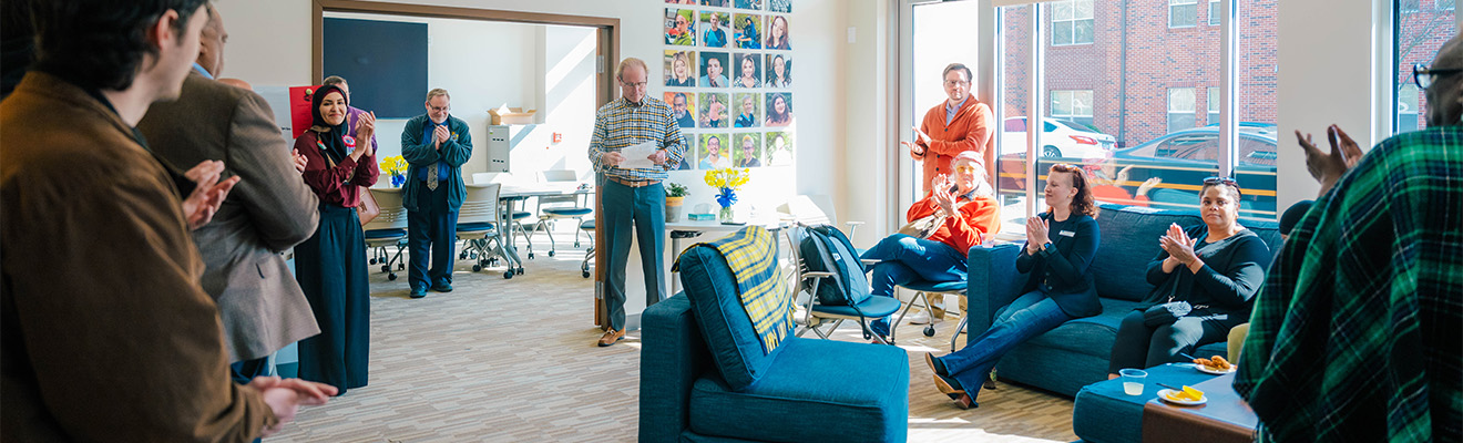 A group of people sit on couches or stand in a room and listen to a man speak.