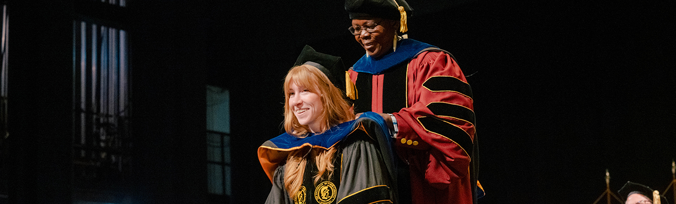 A graduating student, in academic regalia, stands on a stage while Dr. Sandra Ayoo, wearing red doctoral robes and a black academic cap, places a blue hood over the graduate’s shoulders during a hooding ceremony. The stage is dimly lit, with the two individuals illuminated by spotlights against a dark background.