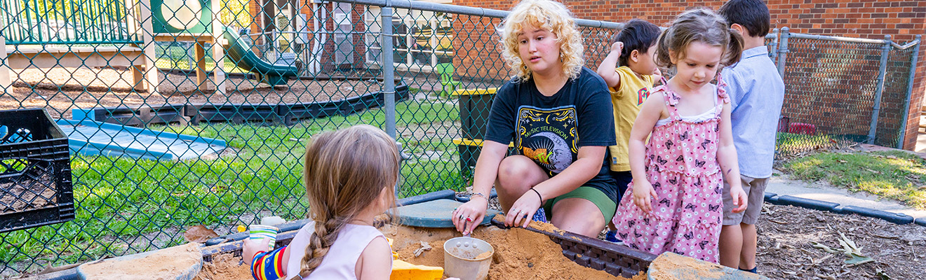 A UNCG student plays in the sandbox with small children.