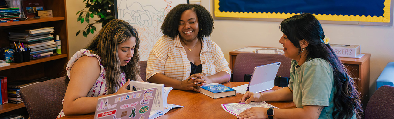 Three students study with laptops and textbooks on a table.