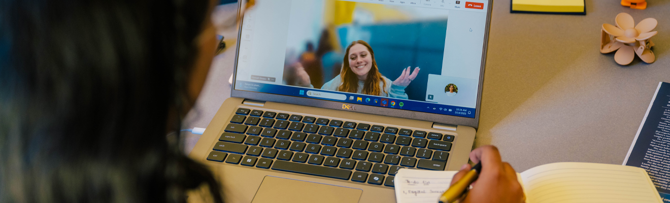 Closeup picture of the back of a student's head who is writing down notes while staring at a video call on a laptop.