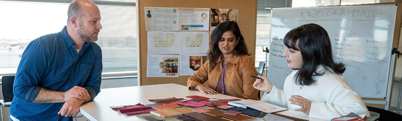 A student and two faculty members stand around a table discussing fabric samples