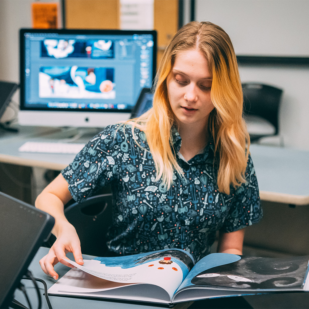 Woman turns pages of a storybook, copies of its illustrations on a computer behind her.