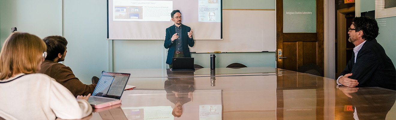 Man stands at head of a conference table in front of a screen with 3 professionals seated at the table and looking on.